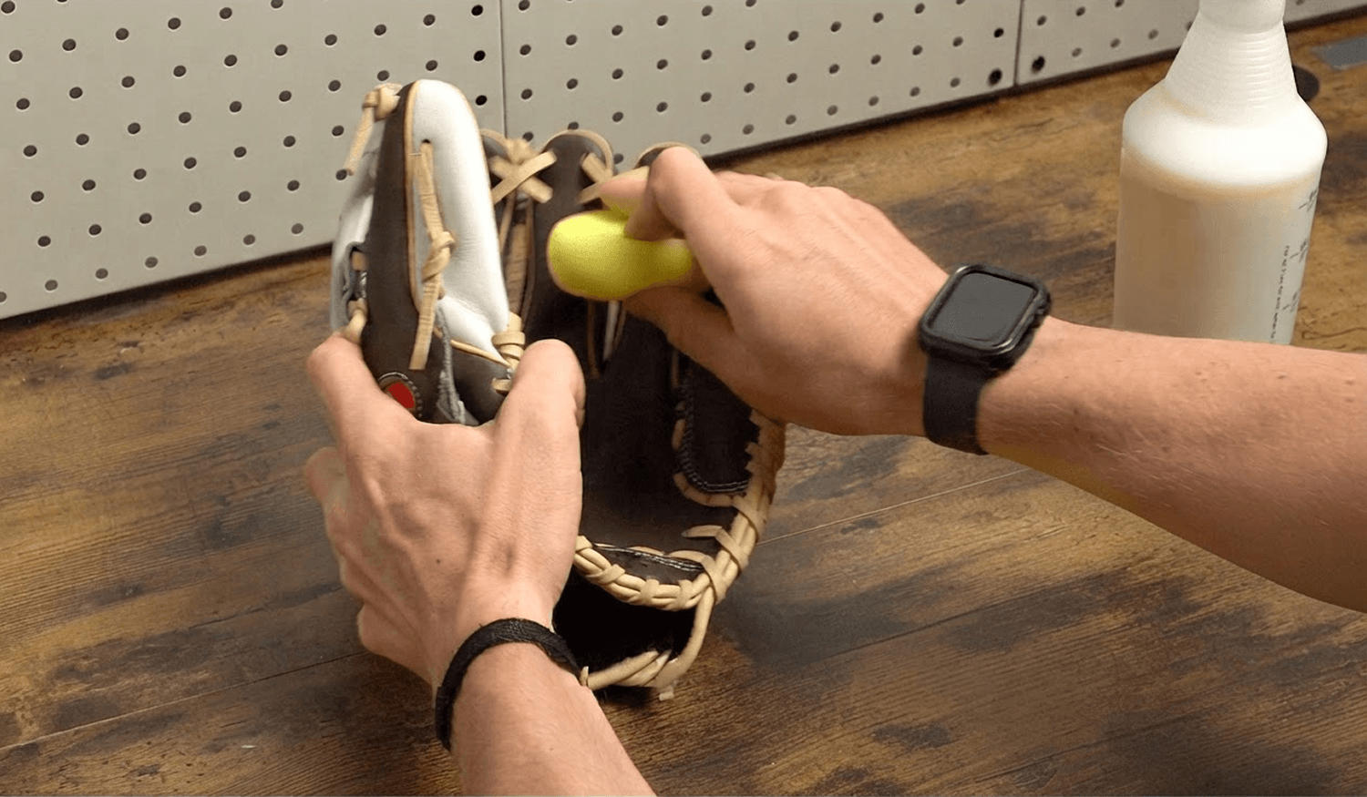 Hands apply a yellow applicator to a baseball glove. A white bottle is visible in the background.