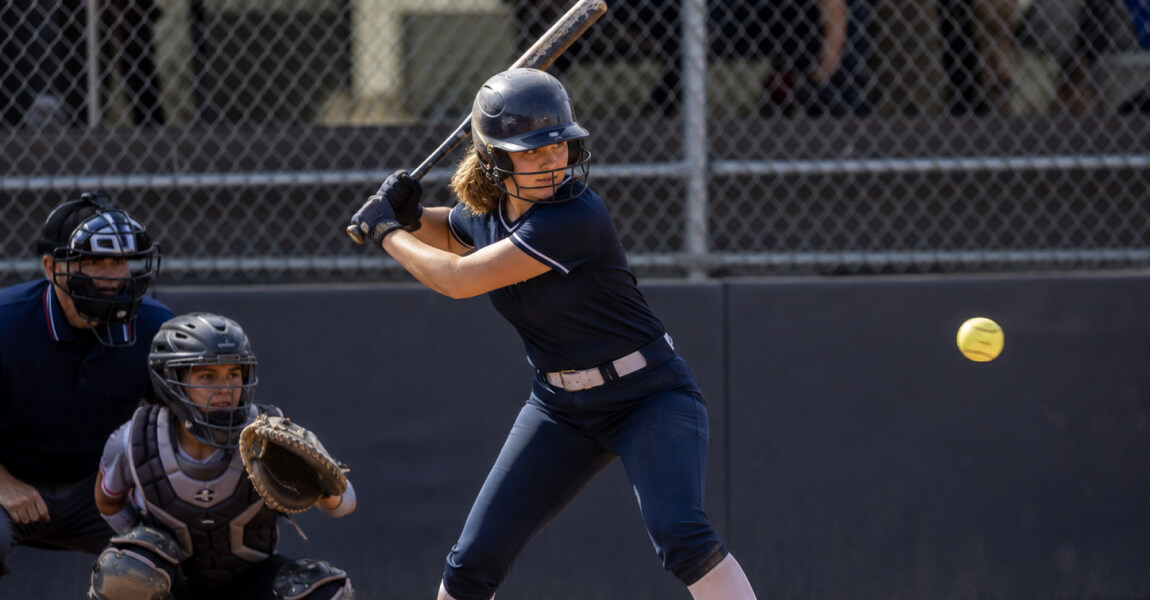 A softball batter swings at a pitch. A catcher and umpire are behind her.