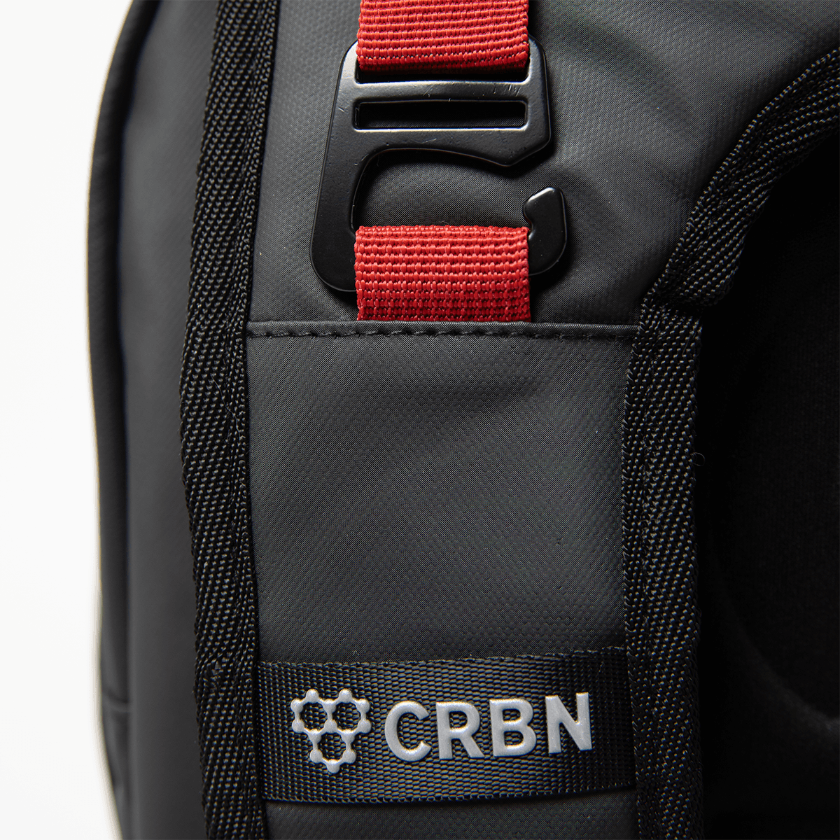 Close-up of a black bag shows a red strap with a black buckle, and a silver "CRBN" logo.
