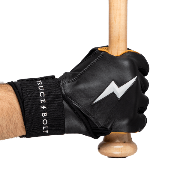 Close-up of a hand wearing a black batting glove with a white lightning bolt logo, holding a wooden baseball bat, and a black wrist strap reading "BRUCE BOLT".
