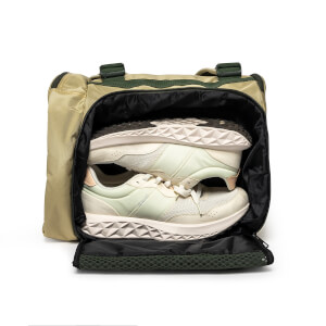 Front view of an open tan duffel bag showing a pair of light cream sneakers with textured soles tucked into a dedicated zippered compartment.