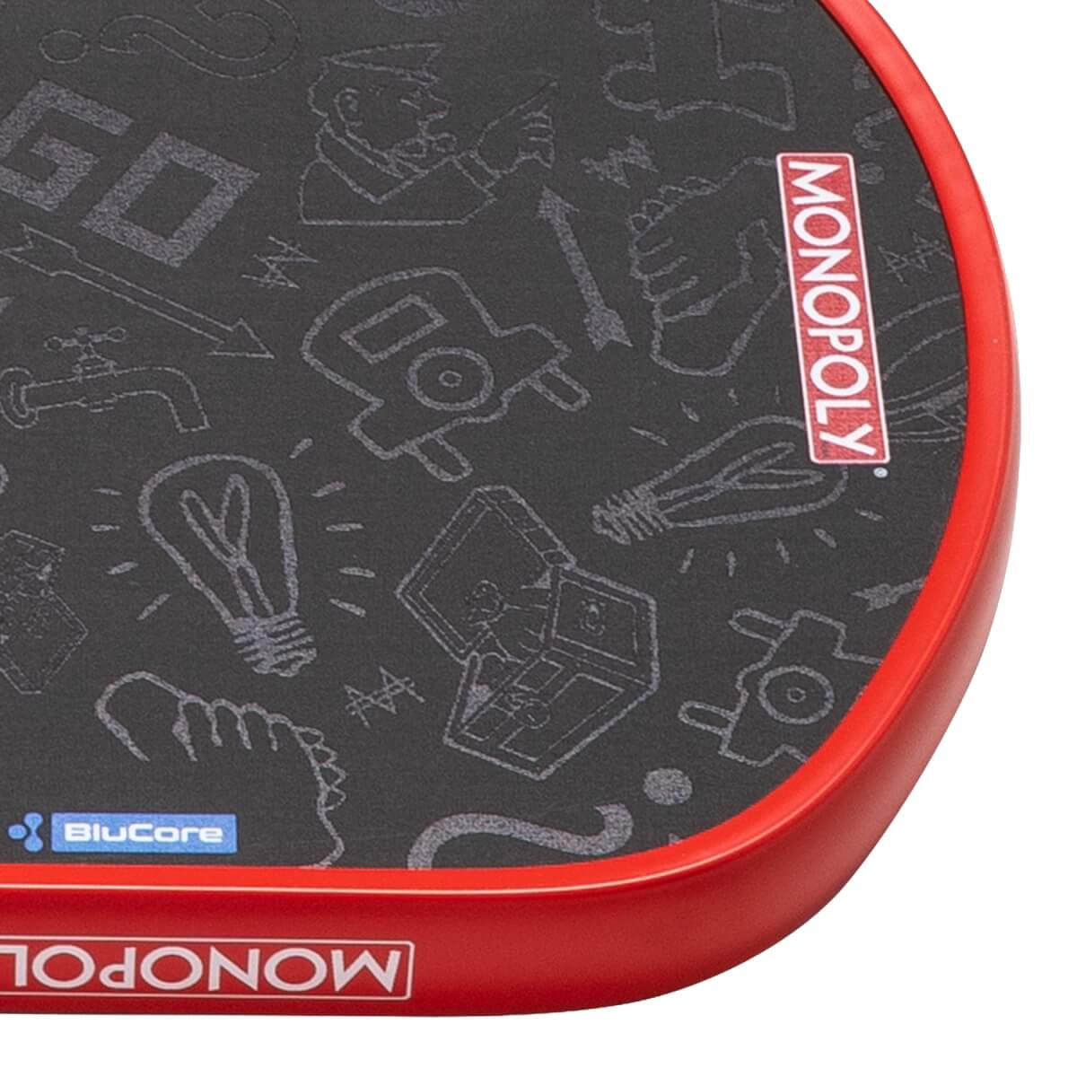Close-up of a dark gray pickleball paddle with Monopoly illustrations, a red edge guard, and MONOPOLY text.