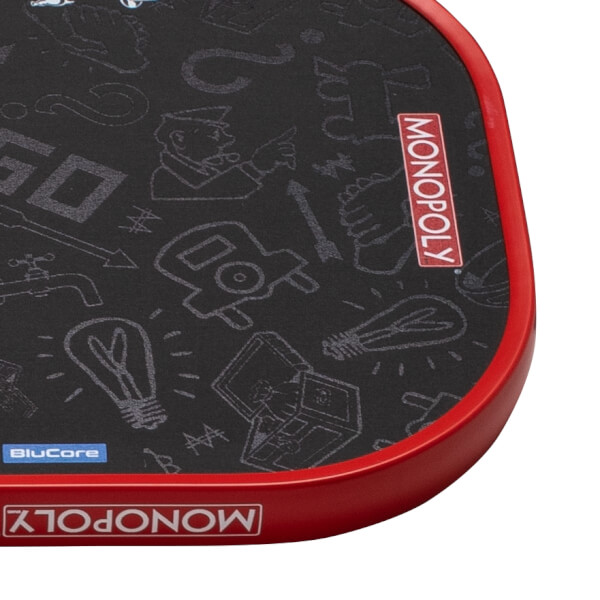 Close-up of a black pickleball paddle corner with Monopoly-themed drawings, a red edge guard, and MONOPOLY and BluCore branding.