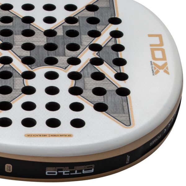 Close-up corner view of the white racket head featuring black holes, gold accents, and the text NOX, AT10 GENIUS, and 12K ALUM XTREM.
