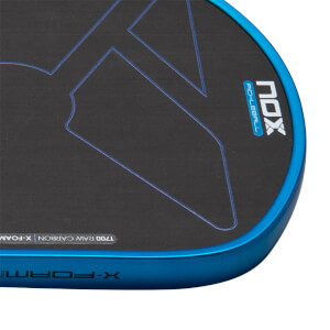 Close-up corner of the paddle featuring the black T700 Raw Carbon face with the blue Nox Pickleball logo and a blue edge guard with X-Foam branding.