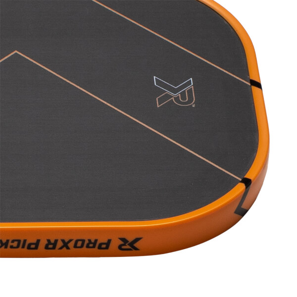 Close-up of a black pickleball paddles top corner with an orange edge guard, diagonal lines, and PROXR PICK text.