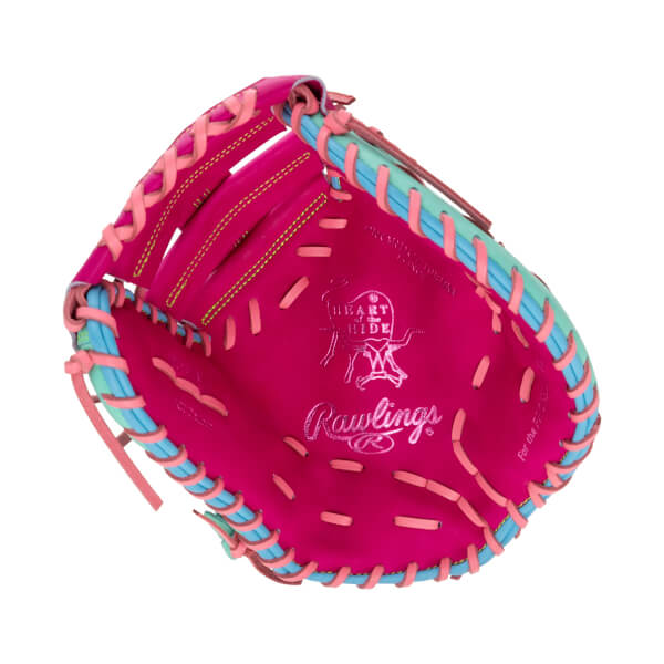 Front palm view of a pink first base mitt with multicolored laces, featuring silver "HEART of the HIDE" and "Rawlings" logos stamped on the leather.