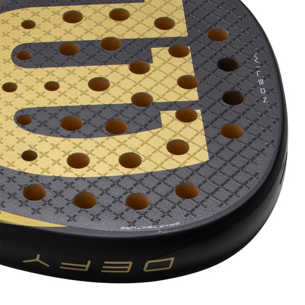 Close-up, angled view of a black and gold padel racket with a large gold W logo, circular holes, and WILSON text.