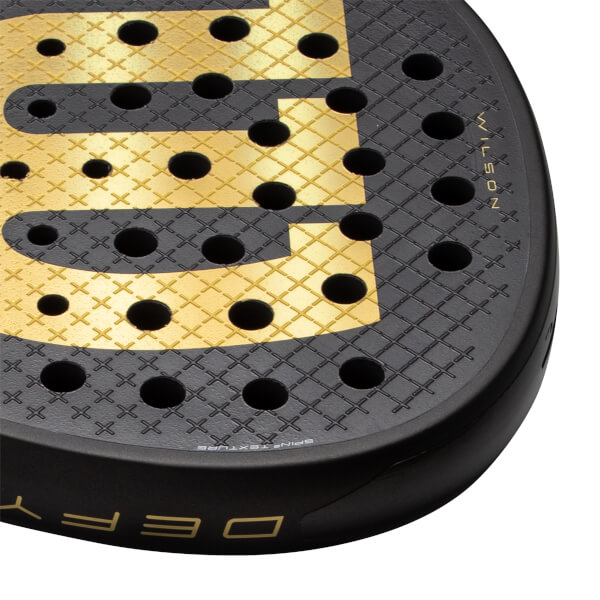 Close-up of a black and gold padel racket with a textured face, round holes, and WILSON and DEFY branding.